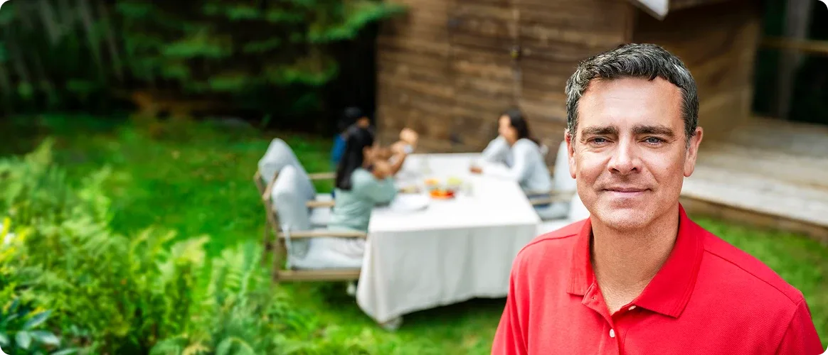 A man smiling and standing outdoors and people are sitting at a table in the background